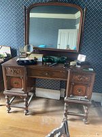 Full view of antique vanity table with large attached mirror and various personal items on top (not included). Visible wooden carvings on drawers and turned legs.