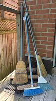 Four assorted household brooms propped against brick wall on wooden deck.