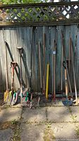 Full view of all garden tools leaning against fence, showing assortment of shovels, rakes, and hand tools