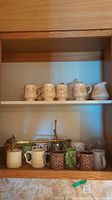 Two shelves in a wooden cabinet with various ceramic and glass mugs arranged in rows, showing different styles and colors.