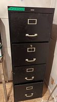 Front view of black metal 4-drawer file cabinet with metal handles and label holders, showing slight wear and keys in lock.