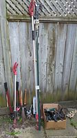 Wide view showing multiple long-handled garden tools including pruners and pole saws leaning against a wooden fence with a box at their base containing smaller tools.