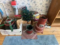 Green metal Christmas tree stand and base seen from above in box, tangled holiday string lights on green wiring nearby, red decorative heater-shaped item seen on side.