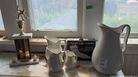 Photo showing three ironstone pitchers, a large trophy with a figurine on top, and a boxed bath cushion on a windowsill.