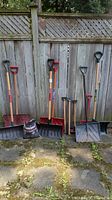 Wide view of snow removal accessories against a wooden fence showing multiple shovels, scrapers, and a bag of salt on the ground.