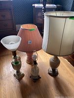 Photo showing four table lamps arranged on wood floor near furniture. Three lamps have shades; one lamp has exposed light bulb and no shade. Ceramic style bases in neutral colors.