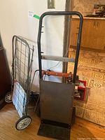Black metal hand truck dolly with solid footplate and folding wheeled cart in background.
