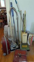 Photo showing three cleaning appliances: red Kenmore 7010 vacuum, white Beaumont vacuum with cloth bag, and green Bissell Foamatic carpet shampooer with wooden handle.