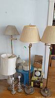 Photo showing five vintage floor lamps with brass/gold-tone metal bases and pleated fabric shades, plus additional ceiling light globes and boxed light kit on floor around a blue stool.