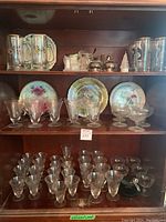 Shelf with decorative plates with floral and bird designs, etched stemware glasses and goblets, clear beer mugs with gold accents.