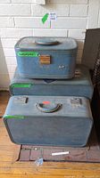 Three vintage blue travel cases stacked: small train case on top with monogram M.L.B., medium suitcase below, largest suitcase at bottom