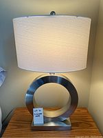 Side view of one silver circular base table lamp with white fabric shade, showing general design and lighting condition.