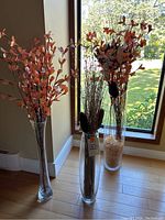Three tall clear glass vases arranged from shortest to tallest with artificial floral arrangements inside, positioned near window with outdoor view.