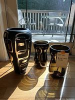 Three black glass vases arranged on a wooden floor near a window, front view showing patterns and general shapes.