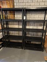 Front view of two black plastic storage shelf units side by side in basement with concrete block wall background.