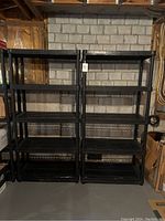 Front view of two black plastic shelving units against a concrete block wall in basement, showing four slatted shelves per unit.