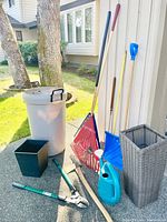 Overall view of Rubbermaid garbage can, planters, rakes, snow shovel, watering can, axe, and hedge trimmers outdoors.