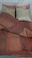 King comforter, two beige decorative cushions with leaf embroidery, two pinkish-brown pillow shams, and folded bed skirt arranged on bed.