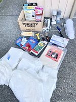 Full lot overview showing paint brushes, rollers, spray paint, vinyl gloves, drop sheets, sanding discs, scraping and sanding tools arranged on concrete.