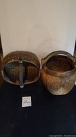Front view of two woven baskets with handles placed upright against a white wall on a black surface.