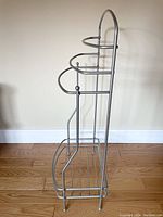 Side view of silver metal towel rack showing three horizontal towel bars and bottom storage rack, standing on wood floor against wall.