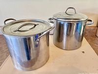 Two stainless steel pots with lids, side by side on a counter under natural light showing entire pots including handles and lids.