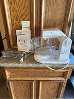 Sunbeam Mixmaster mixer with glass bowl, beaters, and instruction book displayed on countertop against wood cabinetry.