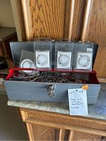 Photo of metal tool box with red lid containing assorted hand tools and hardware. Four mechanical timers in plastic packaging displayed on top of the box.