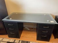 Full front view of the black writing desk showing glass top and six drawers, sitting on wood floor.