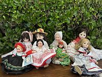 Eight vintage porcelain dolls displayed seated on a wooden surface against greenery background, dressed in various traditional costumes.
