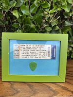 Framed ticket from Slayer/Megadeth American Carnage Tour with guitar pick, front view showing ticket details and green frame.