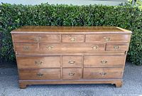 Front view of the wooden dresser showing 12 drawers and brass handles against a hedge background.