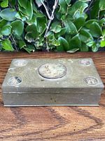 Top and angled view of vintage Chinese brass trinket box showing etched design, coin-like corner medallions with Chinese characters, and central carved medallion.