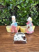 Full lot showing two Dutch children planters and a cat planter on wooden table with green hedge background.