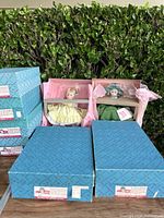 Two Madam Alexander dolls partially visible in original blue boxes with pink tissue paper inside, one dressed in yellow and one in green.