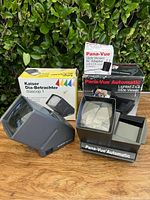 Photo showing both slide viewers displayed with their original boxes and the AC adapter box in the background, set on a wooden surface with green foliage background.