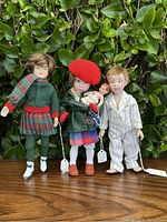 Three vintage Norman Rockwell dolls posed in front of leafy background. Left doll in green knit sweater and plaid skirt, middle doll with red beret and green coat holding accessories, right doll male in white striped pajamas.