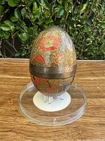 Front view of vintage enameled brass egg on clear plastic round stand showing floral pattern with red and orange flowers and central brass band.