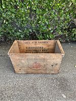 Front and inside views of a rectangular vintage wooden crate marked George Lueders & Co, San Francisco California, with weathered patina and company logo, placed outdoors on pavement with green foliage in the background.