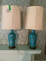 Pair of vintage turquoise ceramic table lamps with gold accents and cream fabric shades, one shade noticeably damaged, on a mantelpiece with wallpaper background.