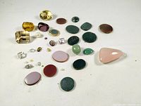 Loose jewelry gemstones arranged on white cloth, showing variety of shapes and colors including yellow, green, pink, red stones, clear crystals, and small pearls.