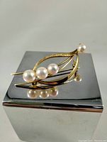 Leaf-shaped gold brooch with five spherical pearls displayed on reflective surface