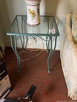 Full view of green painted metal end table with glass top, showing floral ceramic vase on top and floor tiles beneath.