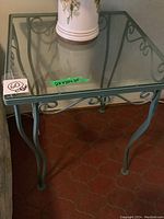 Front and side view of green painted metal end table with glass top showing scroll metalwork and curved legs on tiled floor.