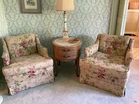 Photo of two floral vintage armchairs flanking a round wooden side table with lamp and decor on top