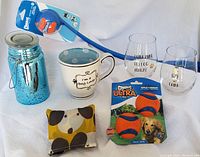 Lot arranged showing all items: Chuckit ball launcher with ball, 2 Chuckit Ultra balls, 2 Mikasa stemless large wine glasses with dog sayings, ceramic dog lover mug, flameless lantern candle in blue mason jar, and folded dog-themed reusable shopping bag.