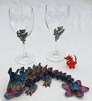 Two clear wine glasses with pewter dragon emblems, small red glass blown dragon, and fully jointed dragon figurine displayed on white cloth.