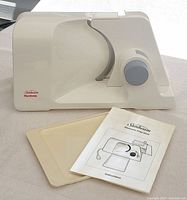 Side view of the Sunbeam electronic food slicer showing the control knob, safety lock, and label with model information along with included platter and instruction booklet.