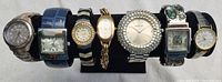 Seven assorted watches on display, various styles and materials visible.