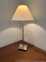 Single cream shade lamp with metal base and pull chain turned on, lit in ambient light, photographed from the side on a wooden table.